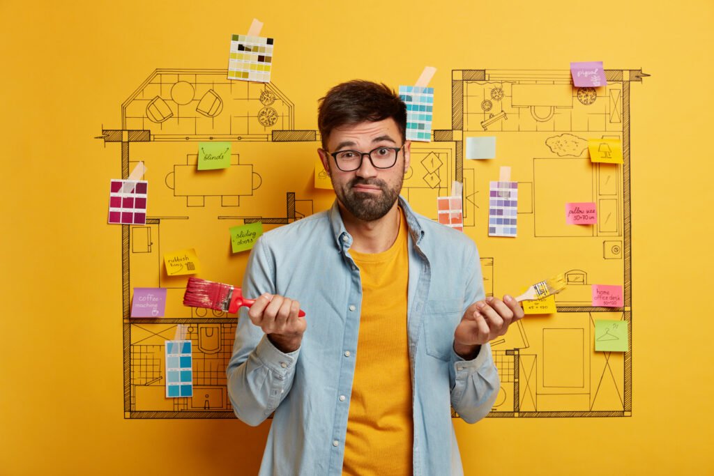 bewildered bearded man holds two paintbrushes, feels hesitant and confused, thinks about designing new apartment, house decoration and renovation. puzzled house painter does repairing at home.