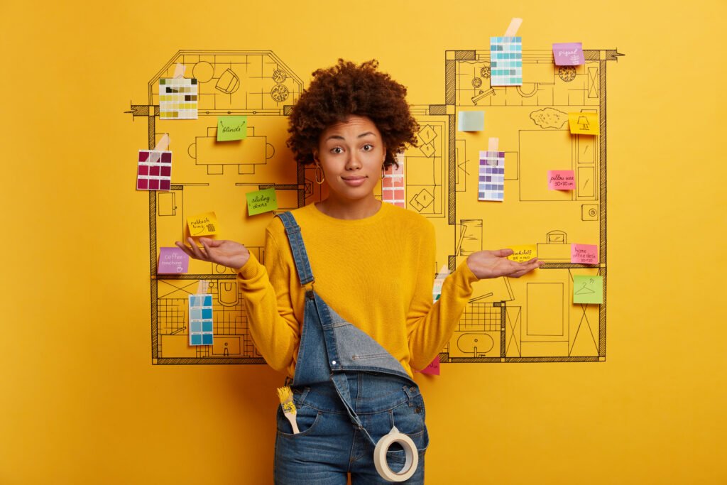 hesitant woman architect poses against drawing sketch, spreads palms sideways, makes decision how to remodel house better, wears casual yellow sweater, denim overalls with paint brush in pocket